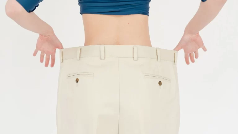 Back view of faceless fit female wearing white oversized trousers standing on white background in light studio after weight loss