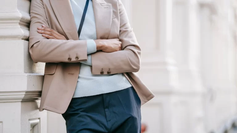 Crop anonymous stylish female manager in smart casual outfit leaning on elegant building wall with folded arms while standing on street on sunny day