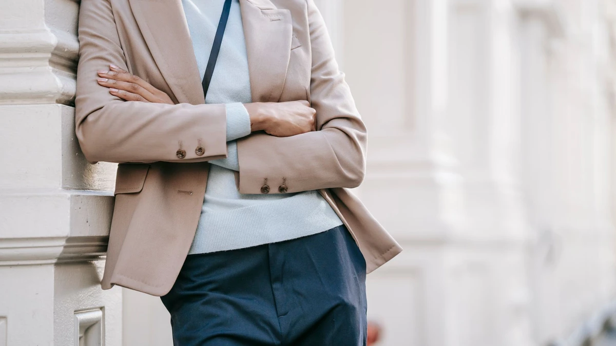 Crop anonymous stylish female manager in smart casual outfit leaning on elegant building wall with folded arms while standing on street on sunny day